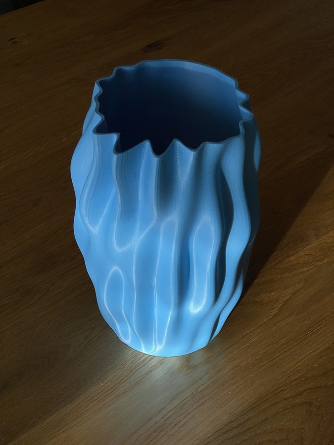 A sky-blue 3D-printed vase seen from above on an oak table, deep vertical ridges down one flank and a jagged sawtooth rim