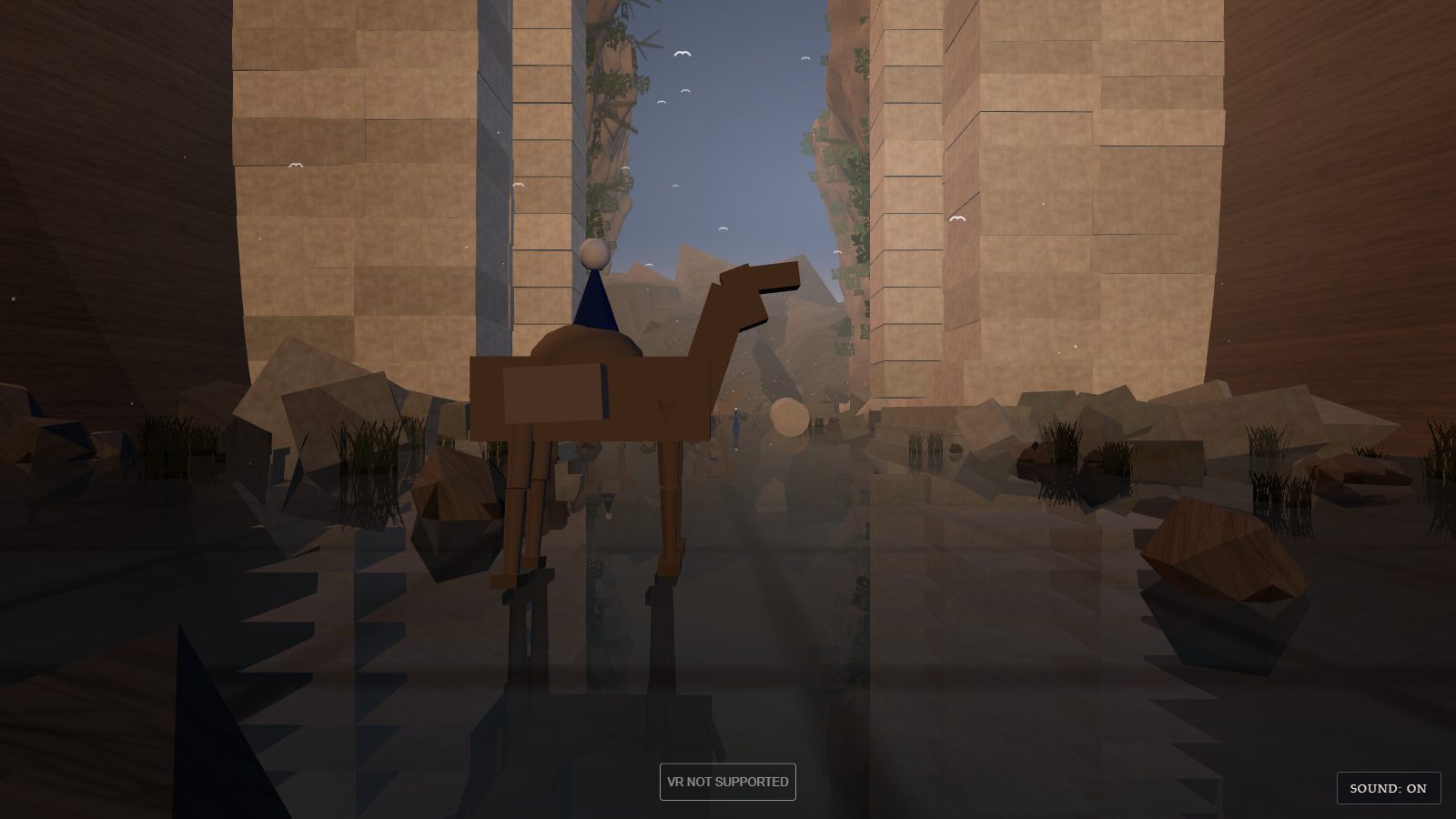 Looking back at the gate from the far side, in daylight blue. The inner faces of the piers are plain sandstone blocks catching the light. A boxy camel with a rider in a blue cone-shaped robe stands in the water in front of the opening, built from simple rectangular blocks. Boulders and reeds line both sides